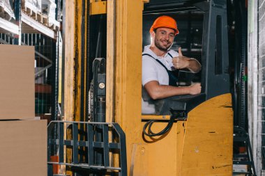 Gülümseyen depo çalışanı, forklift yükleyicisini çalıştırırken başparmağını kaldırıyor ve kameraya bakıyor.