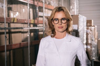 Beyaz önlüklü kendine güvenen depo sahibi depodaki kameraya bakıyor.