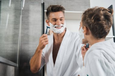 selective focus of happy father with shaving foam on face looking at son in bathroom  clipart