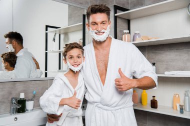 cheerful father and son with shaving foam on faces showing thumbs up in bathroom  clipart