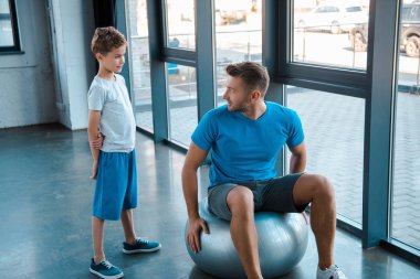 Babasının yanında duran yakışıklı çocuk spor salonunda fitness topu üzerinde çalışıyor. 
