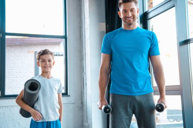 Mutlu bir baba elinde halterlerle yakışıklı bir çocuğun yanında spor paspası tutuyor. 