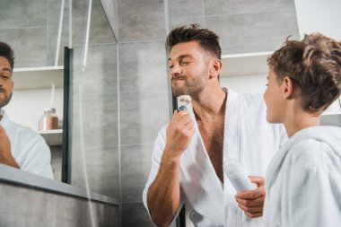selective focus of handsome father holding shaving brush near face while standing near son in bathroom  clipart