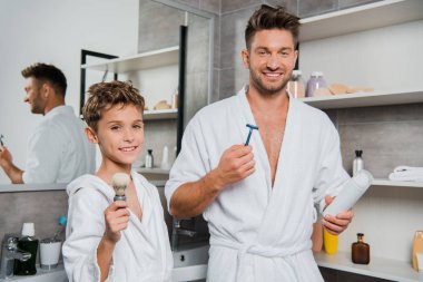 happy father holding razor and can with shaving foam near son with shaving brush  clipart