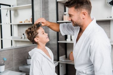 handsome father brushing hair of cute son in bathroom  clipart