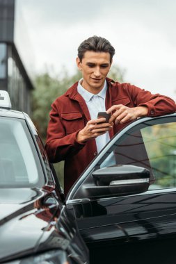 Arabanın yanında akıllı telefon kullanan yakışıklı bir taksi şoförü.