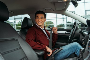 Smiling man holding safety belt in car and looking away clipart