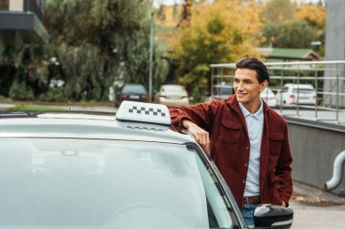 Gülümseyen taksi şoförü arabanın yanına bakıyor.