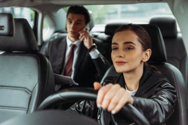 Kadın taksi şoförü dümeni tutarken iş adamı akıllı telefondan konuşuyor.