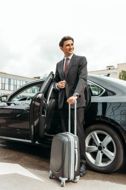 Smiling businessman with suitcase standing by taxi clipart