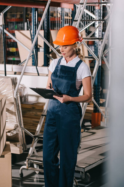 selective focus of workwoman in overalls and helmet standing near construction materials and writing on clipboard