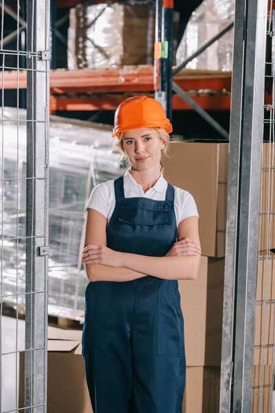 attractive workwoman in overalls and helmet standing with crossed arms and looking at camera