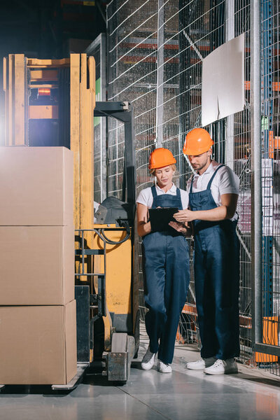 two warehouse workers looking at clipboard while standing near forklift loader