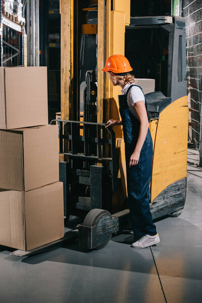 workwoman in overalls and helmet standing near foklift loader in warehouse