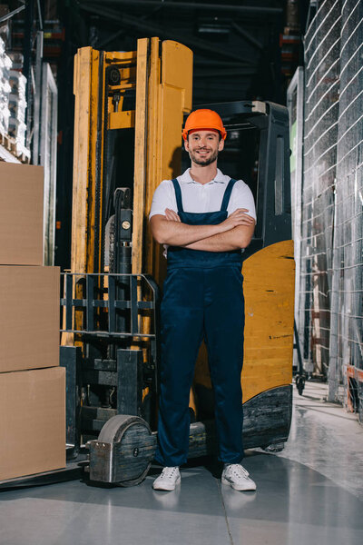 handsome warehouse worker smiling at camera while standing near forklift loader with crossed arms