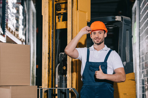 smiling warehouse worker touching helmet and showing thumb up while standing near forklift loader