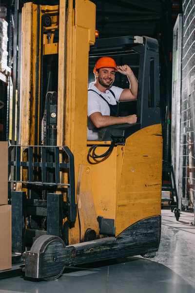 smiling warehouse worker touching helmet and looking at camera while operating forklift loader