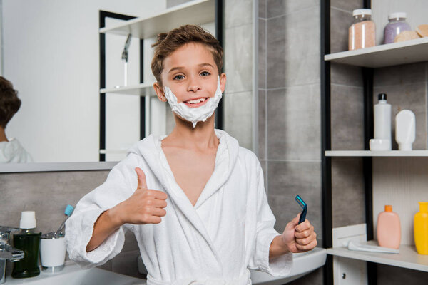happy boy with shaving foam on face holding razor and showing thumb up 
