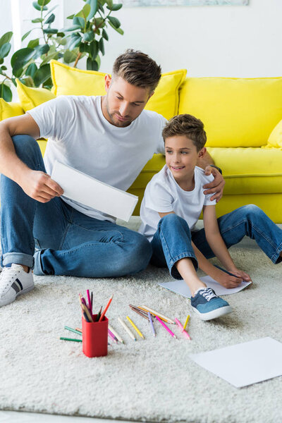happy son sitting on carpet near father colorful pencils and handsome father 
