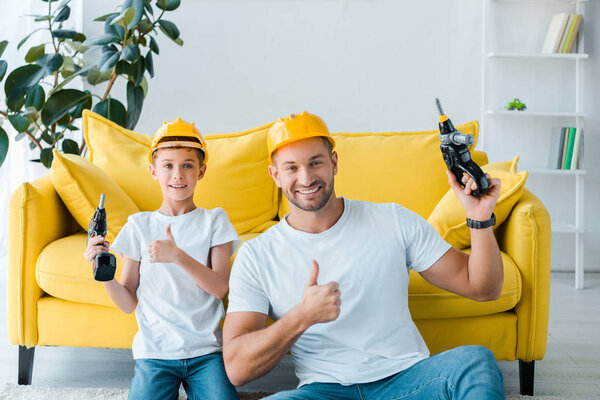 счастливый сын и красивый отец в защитных шлемах показывая большие пальцы вверх и держа молоток сверла
 