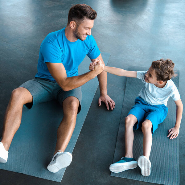 overhead view of cheerful father and son holding hands while sitting on fitness mats 