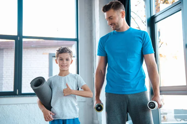 Güzel oğlunun yanında elinde dambıl tutan mutlu bir baba spor salonunda baş parmağını gösteriyor. 
