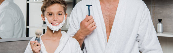 panoramic shot of father and son with shaving foam on faces holding razor and shaving brush 