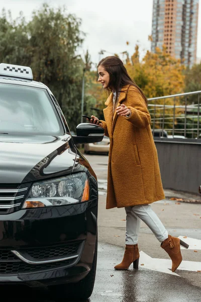 スマートフォンを持って車の横でタクシーの運転手と話す若い女性 — ストック写真