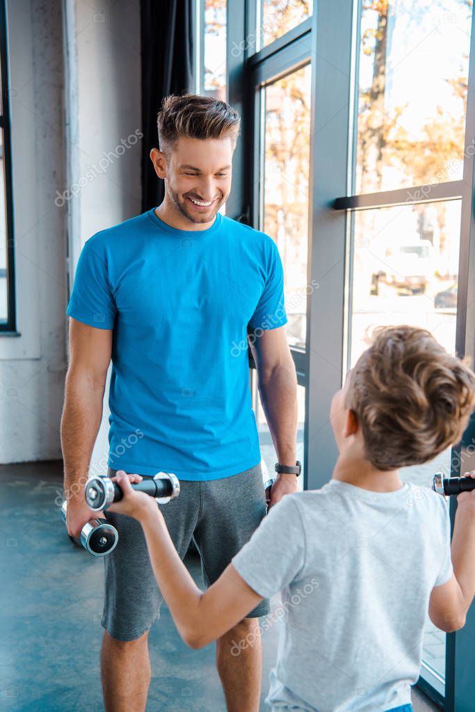 selective focus happy father looking at cute son exercising with dumbbells in gym 
