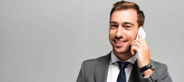 Takım elbiseli yakışıklı ve gülümseyen iş adamının gri renkte izole akıllı telefondan konuşmasının panoramik görüntüsü. 