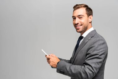 Gülümseyen ve takım elbiseli yakışıklı iş adamı gri üzerinde izole akıllı telefon kullanıyor. 