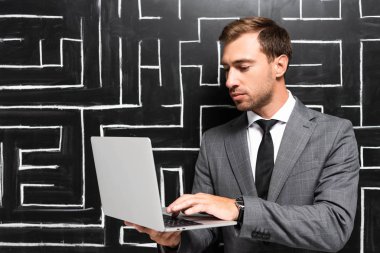 handsome businessman in suit standing near labyrinth and using laptop  clipart