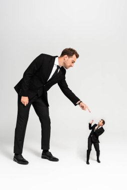 angry businessman in suit pointing with finger at frightened marionette on grey background  clipart