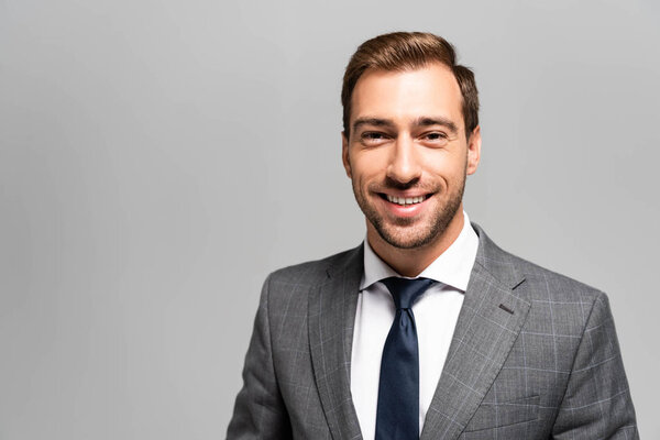 handsome and smiling businessman in suit looking at camera isolated on grey 