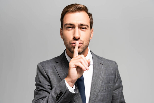 handsome businessman in suit showing shh sign isolated on grey 