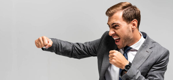 panoramic shot of angry and handsome businessman in suit punching isolated on grey 