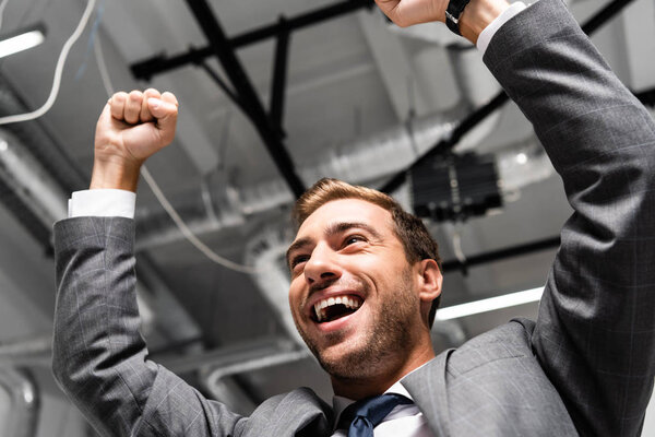 low angle view of handsome and smiling businessman in suit showing yes gesture in office 