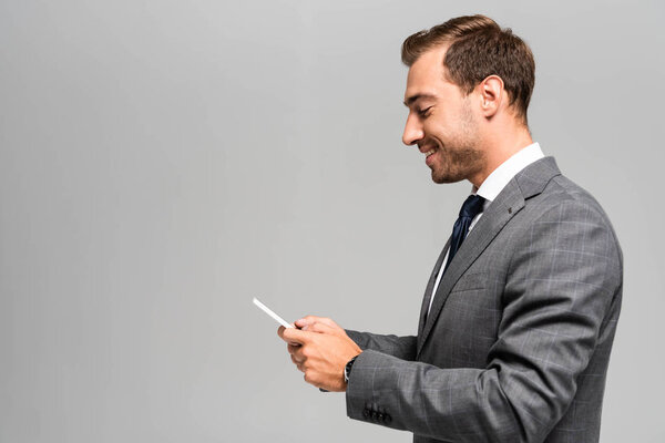 side view of smiling and handsome businessman in suit using smartphone isolated on grey 