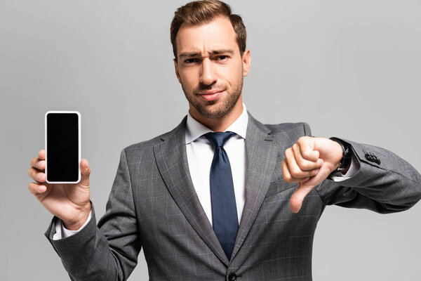sad businessman in suit holding smartphone and showing dislike isolated on grey 