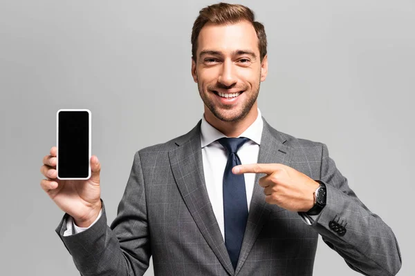 Pohledný Usmívající Podnikatel Obleku Ukazuje Prstem Smartphone Izolované Šedé — Stock fotografie