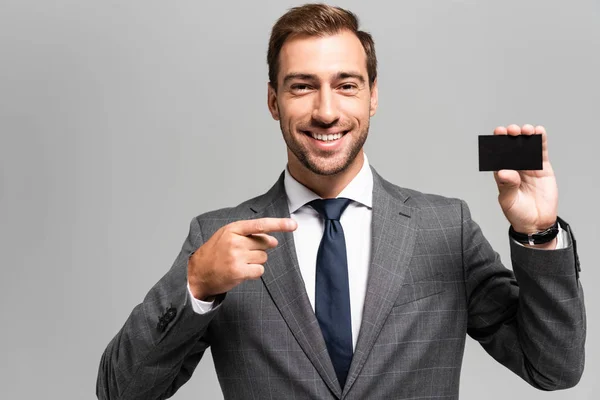 Uśmiechnięty Biznesmen Garniturze Wskazujący Palcem Wizytówkę Odizolowaną Szaro — Zdjęcie stockowe