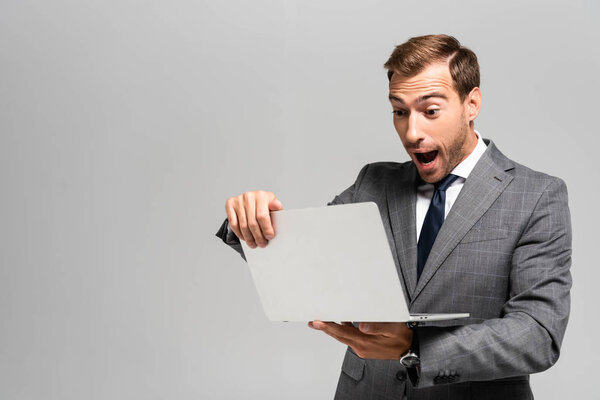 handsome and shocked businessman in suit holding laptop isolated on grey 