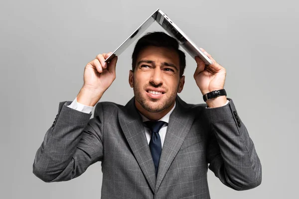 handsome and frightened businessman in suit holding laptop above head isolated on grey