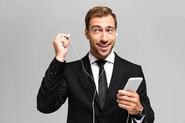 Przystojny Uśmiechnięty Biznesmen Garniturze Słuchawkami Przy Użyciu Smartfona Odizolowanego Szaro — Zdjęcie stockowe