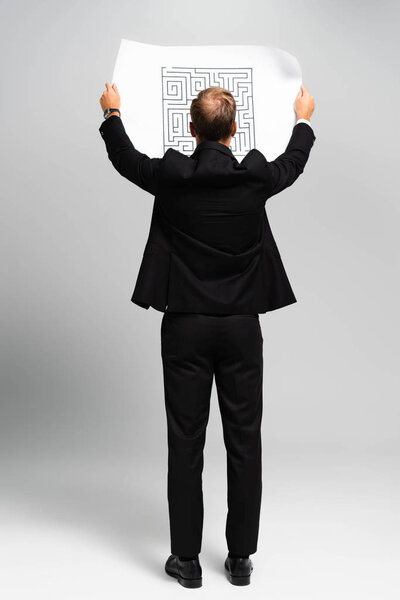 back view of businessman in suit looking at paper with labyrinth on grey background 