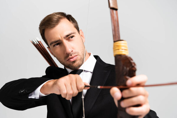 selective focus of handsome businessman in suit holding bow and shooting with arrow isolated on grey