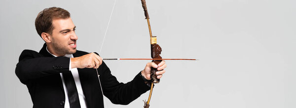 panoramic shot of angry businessman in suit holding bow and shooting with arrow isolated on grey