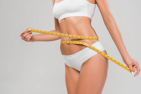 partial view of beautiful slim woman in underwear holding measuring tape on waist isolated on grey
