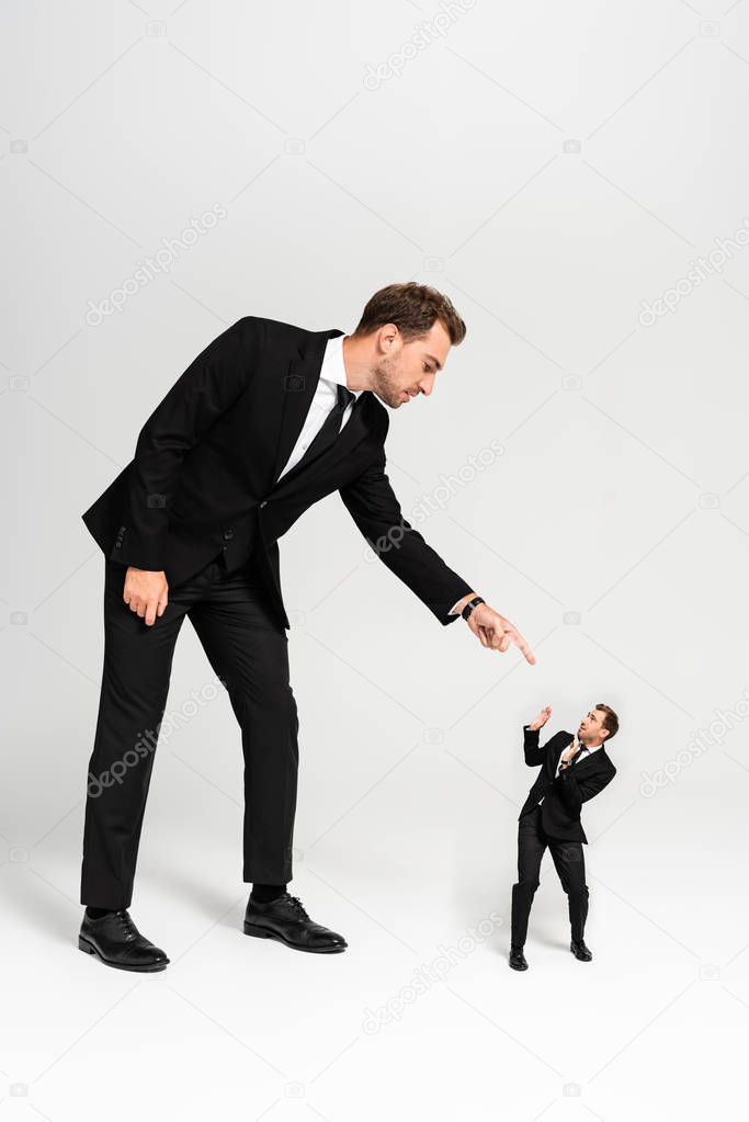 angry businessman in suit pointing with finger at frightened marionette on grey background 