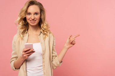 Akıllı telefonu tutan ve pembe üzerine izole edilmiş parmağıyla işaret eden gülümseyen kadın.